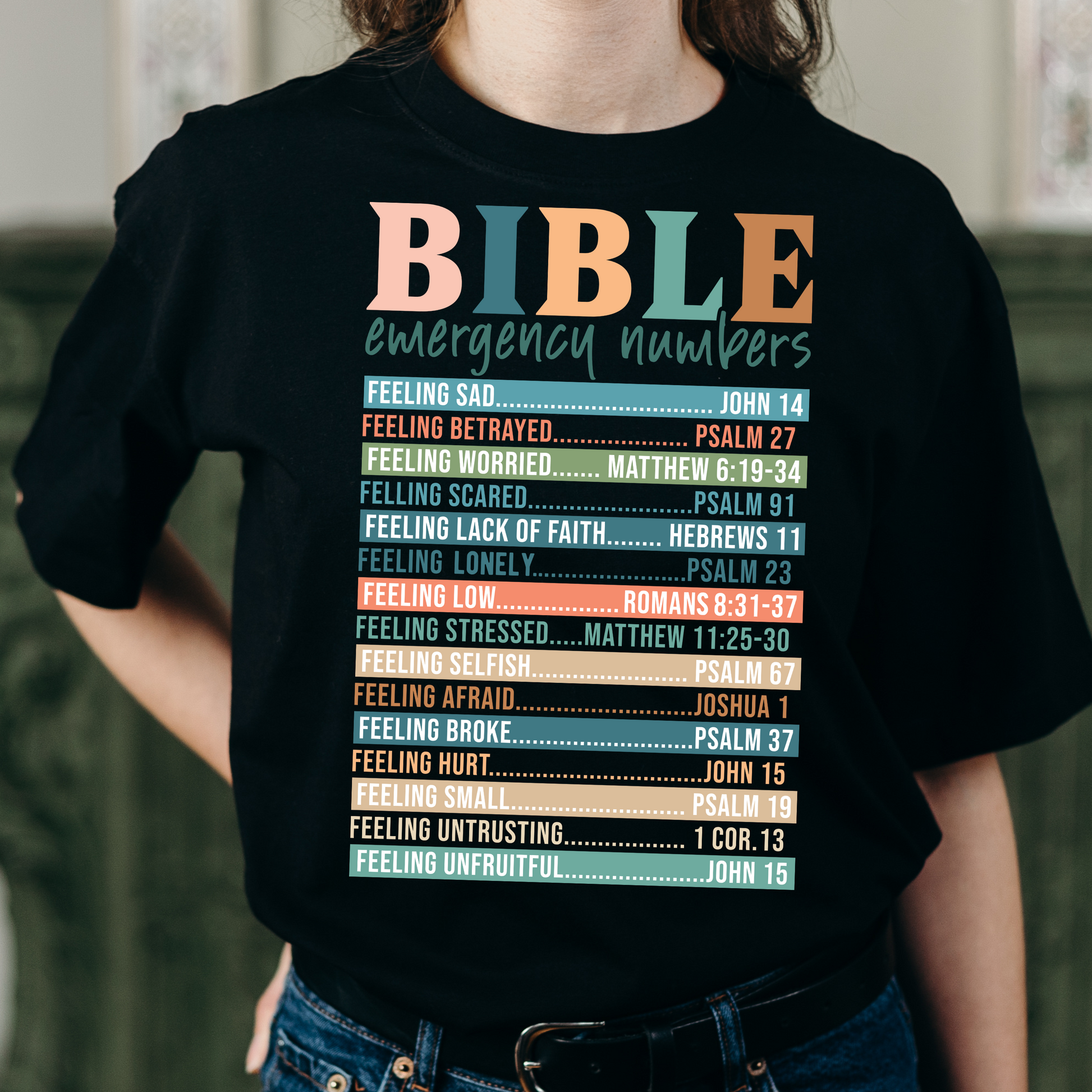 A person wearing a white long-sleeved T-shirt with 'BIBLE emergency numbers' printed in colorful letters, followed by lists of Bible verses.