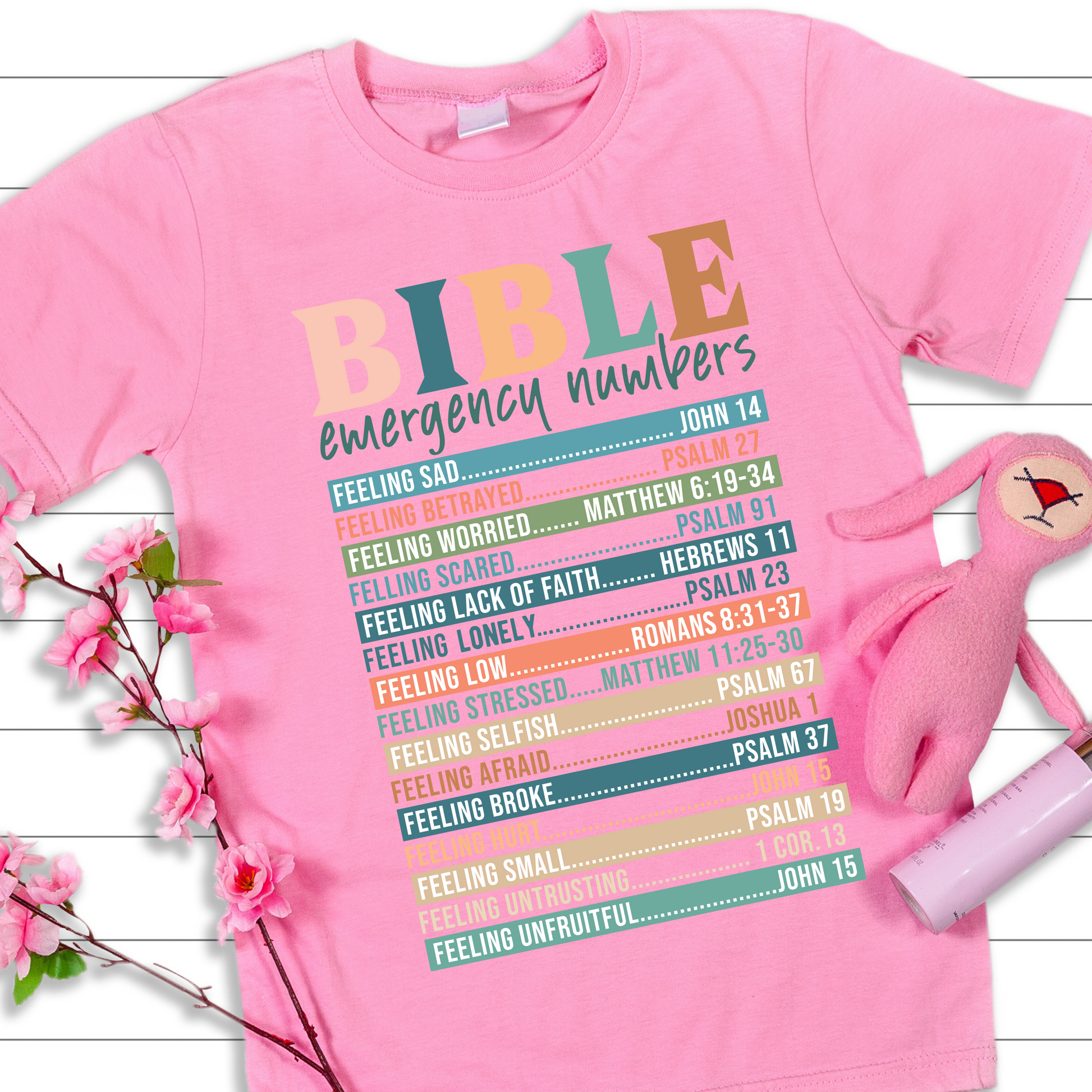 A person wearing a white long-sleeved T-shirt with 'BIBLE emergency numbers' printed in colorful letters, followed by lists of Bible verses.