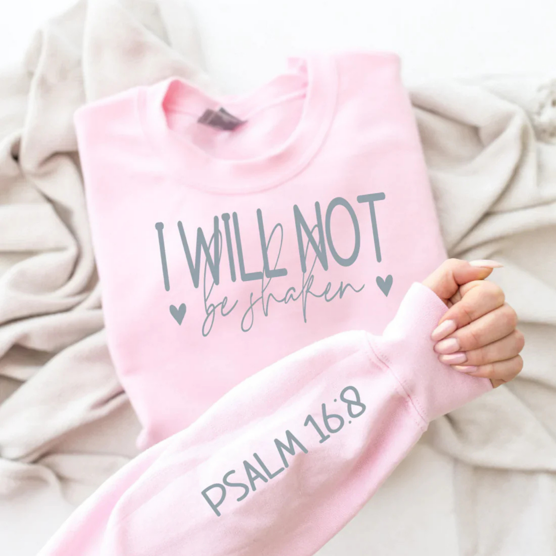 A pink long sleeve t-shirt with the text 'I WILL NOT be shaken' and the reference 'PSALM 16:8' printed on it, held by a person against a neutral background.
